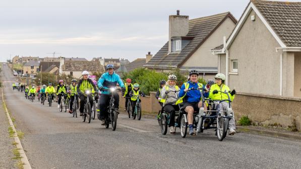 A big line of people are cycling along a quiet village road. There are different kinds of bikes, with non-standard bikes represented. There is a mix of children and adults