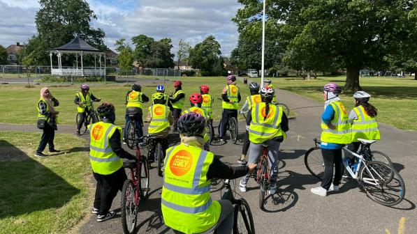 A group of cyclists wearing high-visibility vests gathered in a park, preparing for a group ride under a sunny sky with scattered clouds. A bandstand and lush green trees are visible in the background.