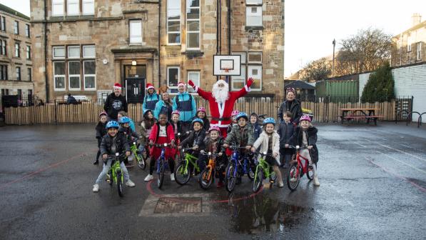 Santa Claus poses with a group of smiling children on brightly colored bicycles in a school playground, with staff members and a historic school building in the background. The festive atmosphere is enhanced by Santa hats worn by some adults.