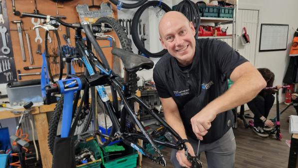 A mechanic at Trackside Bikes smiling while working on a bicycle in a well-equipped workshop. The background features tools, spare bike parts, and wheels neatly organised on the walls and shelves.