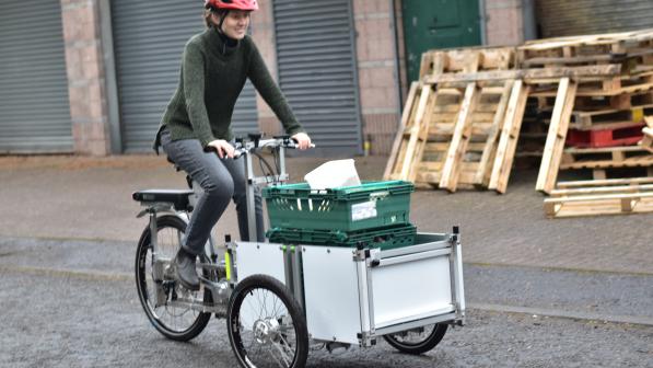 A person cycling a cargo trike loaded with green trays, used for transporting food and materials, on a street lined with wooden pallets and industrial shutters in Craigmillar.