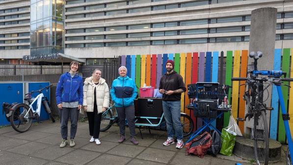 Four people are standing next to their e-cargo bike, tools, and bikes at Glasgow Royal Infirmary. They are in front of a colourful fence, with the hospital building visible in the background.