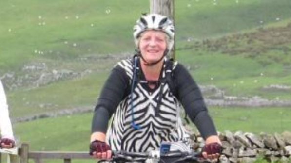 Tina, a smiling woman wearing a zebra-print tunic over cycling gear, rides a mountain bike along a rural road. She wears a helmet and gloves, with rolling green hills and stone walls in the background.