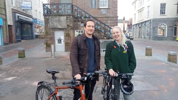 Two NHS medical students standing with their bicycles in front of a historic building in Dumfries town center. The students, smiling and dressed in casual attire, appear ready to ride, with helmets and bikes prominently displayed in the foreground.