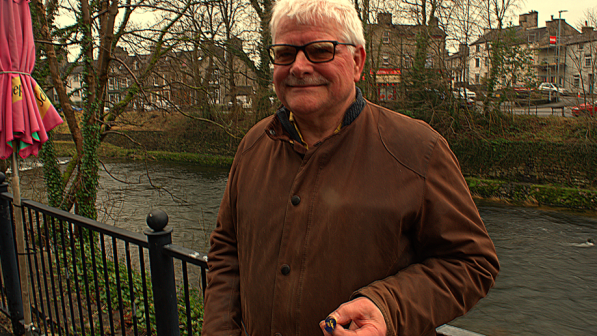 A man in a brown jacket is holding a badge. He's outside in what looks like a pub or cafe garden next to a river