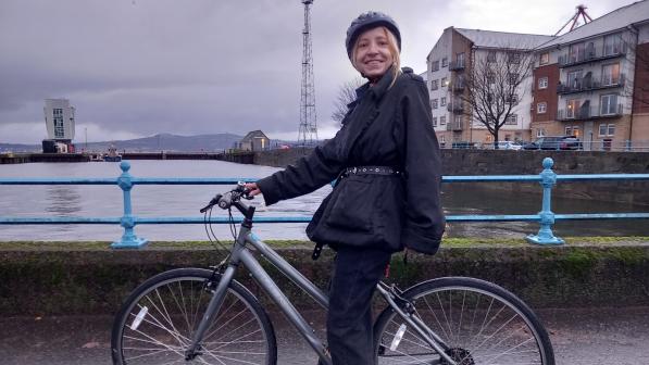 A smiling woman wearing a helmet and dark clothing stands next to her silver bicycle on a waterfront path. Behind her, a blue railing separates the path from the calm water, with an industrial dock, a tall metal tower, and modern apartment buildings in the background. The overcast sky suggests a cool or rainy day.