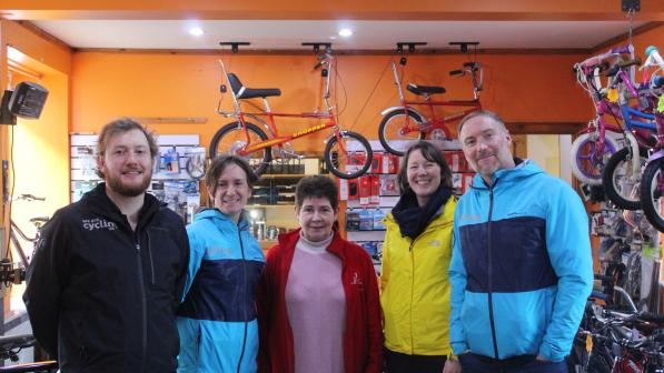 A group of five people stands together inside a bicycle shop with an orange wall, smiling at the camera. Two of them wear matching blue jackets, while another wears a yellow jacket, and a woman in a red cardigan stands in the center. Behind them, vintage red Chopper bikes hang from the ceiling, with various cycling accessories and tools displayed on the walls. The setting reflects a community cycling initiative, likely part of Cycling UK’s work in Shetland.