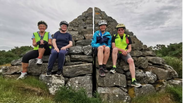 Four cyclists wearing helmets and outdoor cycling gear sit on a historic stone cairn with a visible crack down the middle. They are smiling and appear relaxed, enjoying a break during their ride. The background features an overcast sky and lush greenery.