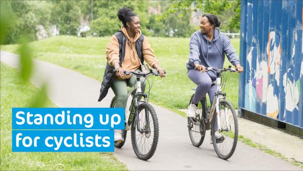 Two women cycling on a path through a park with green fields either side of the path