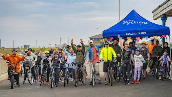 A diverse group of people on bicycles pose and wave in front of a blue Cycling UK tent at a Big Bike Revival event. The group includes children and adults wearing helmets, with colourful bunting decorating the tent.