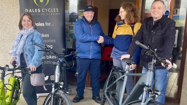 Four people stand outside a building with e-bikes, smiling. A banner for "Dales Cycles" is visible in the background. Two individuals in the center shake hands, marking the delivery of new e-bikes.