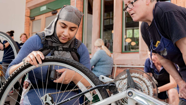 A woman wearing a headscarf and backpack carefully inspects the rear wheel of a bicycle, while a bike mechanic in glasses and a blue apron leans in to provide guidance. 
