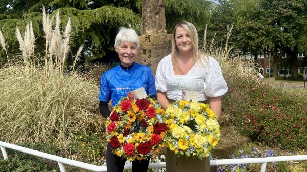 Two women stand smiling in front of the National Cyclists’ Memorial at Meriden, each holding a colourful floral wreath. One wears a blue Cycling UK jersey, the other a white blouse and khaki skirt. Several wreaths are laid on the grass behind them.