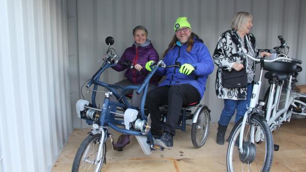 Three smiling adults try out adaptive and electric bikes inside a storage container. Two are riding a side-by-side tandem trike, while a third person stands next to an e-bike.