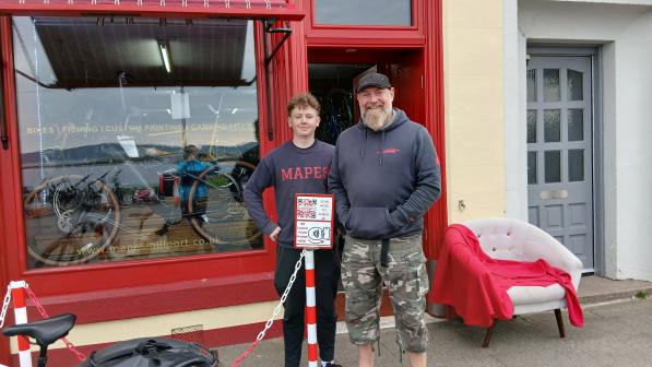 Scott Ferris and another staff member stand outside Mapes of Millport, a bike shop with red trim and a display window showing bikes inside. A bike with panniers is parked in the foreground.