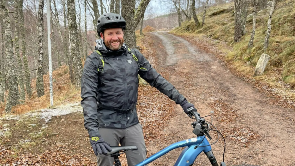 Robbie Hope stands smiling with his blue e-bike on a forest path surrounded by leafless trees. He is wearing a black helmet, waterproof jacket, and gloves. The winding trail behind him is lined with moss and fallen leaves, suggesting a chilly but scenic woodland ride.