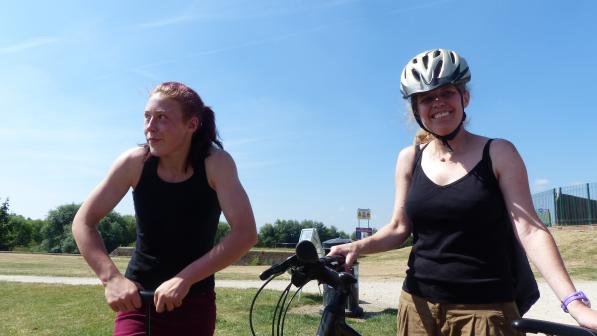 Two smiling women in sleeveless tops enjoy a sunny day outdoors. One is wearing a bike helmet and standing beside a bicycle, while the other stands beside her with a relaxed posture. A dirt path and grassy parkland are visible in the background.