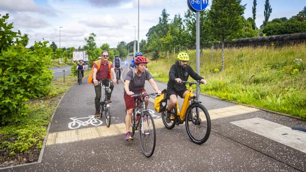 A group of adults riding bikes along a shared cycle and pedestrian path in a green, suburban area. The lead riders include a woman on a hybrid bike and a man on a yellow e-bike. Everyone is wearing helmets and casual clothing, enjoying the ride under a partly cloudy sky.