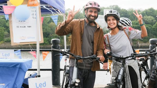 Two smiling cyclists wearing helmets flash peace signs while standing with their bikes at a Big Bike Revival event. Behind them is a riverside tent decorated with colourful bunting and signs offering free led rides.