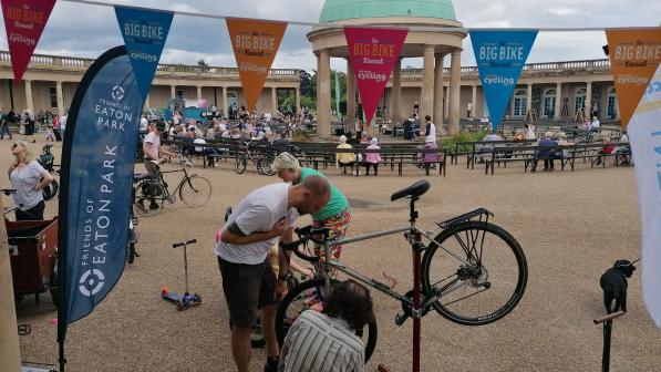A community cycling event takes place in a large open plaza with a domed pavilion in the background. People are seated at benches and walking around. In the foreground, two mechanics work on a bicycle under bunting that reads “Big Bike Revival,” with a “Friends of Eaton Park” flag nearby.