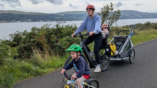A parent rides an e-bike along a waterside path, towing a trailer with a young child, while another child cycles ahead on a small bike. Hills and water are visible in the background.