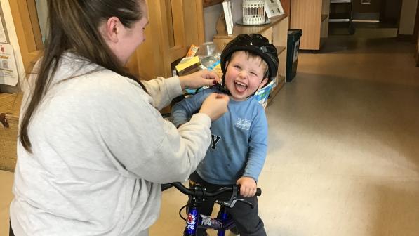 A smiling young child sits on a small bike indoors while an adult helps fasten their helmet. The child looks excited and happy, ready to ride.