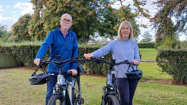 Two adults stand outdoors with their electric bikes on grass, with trees and a blue sky in the background.
