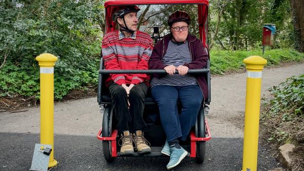 Two adults wearing helmets sit side by side on the front seat of a red adapted cycle, positioned between two yellow bollards on a shared-use path surrounded by trees.