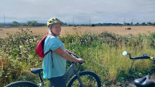 Adult woman in a yellow cycle helmet smiles while standing with a grey bike on a rural path. She wears a turquoise top and red backpack. Behind her are hay bales, hedgerow and pylons under a cloudy sky.