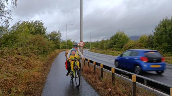 Adult rides a yellow bike on a wet shared path beside a 40-mph road, with a child in a front seat wearing a yellow rainsuit and blue helmet; overcast sky and a car passing.