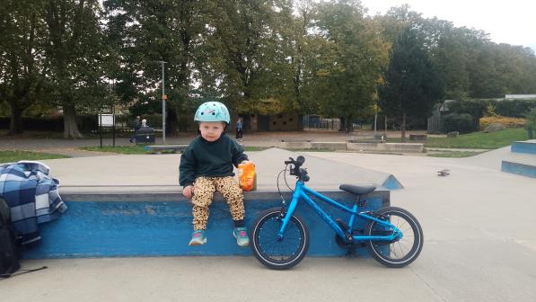 A young child wearing a turquoise helmet sits on the edge of a low skatepark ramp, holding a bag of snacks. Next to them is a small bright blue pedal bike. Trees and other skatepark features are visible in the background.