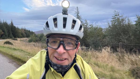 Selfie of a cyclist in a yellow jacket wearing a helmet with a mounted headlight and protective glasses, riding on a rural road beside trees at dawn/dusk under a cloudy sky.