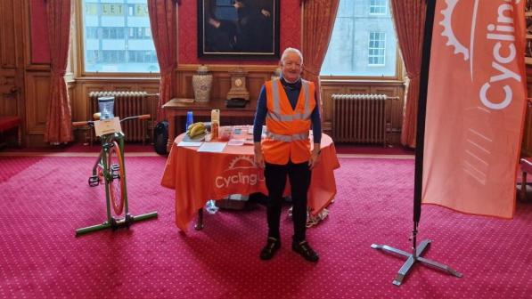 Older adult in a high-vis orange vest stands in a grand, red-carpeted room beside a Cycling UK table and banner, with a static bike display and tall windows.