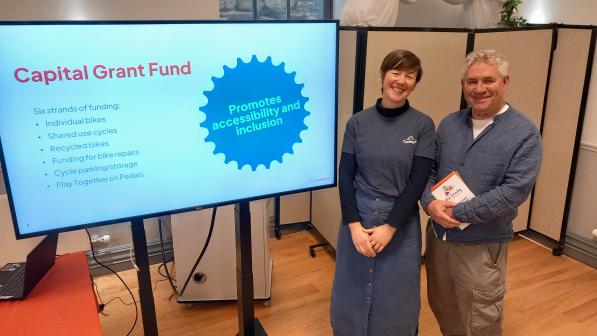 Two adults standing beside a TV screen showing a ‘Capital Grant Fund’ slide with a badge reading ‘Promotes accessibility and inclusion.’ The slide lists funding strands including individual bikes, shared-use cycles, recycled bikes, funding for bike repairs, cycling infrastructure/parking, and Play Together on Pedals. Indoor community venue.