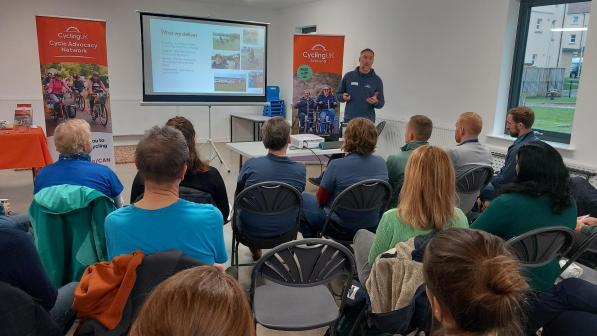 A group of people sit on folding chairs in a bright meeting room, facing a screen where a Cycling UK presentation is being given. A speaker stands beside the screen in front of two Cycling UK Scotland banners, talking to the audience about cycling while slides with photos and text are projected.