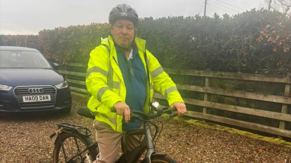 An older man wearing a black helmet and bright yellow hi-vis jacket sits on a dark e-bike on a gravel driveway, with a parked car and wooden fence behind him on an overcast day.