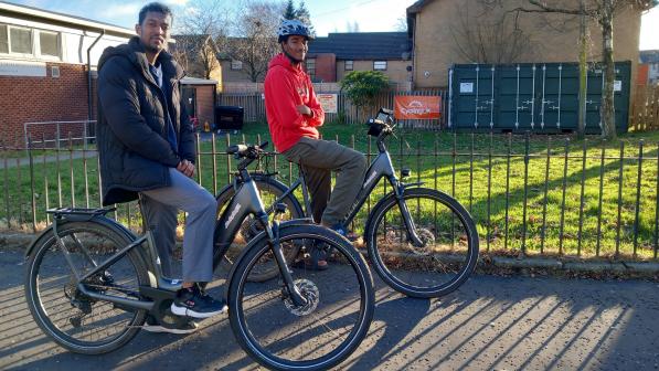 Two college students on e-bikes pause on a residential street in Glasgow, one wearing a red hoodie and helmet, the other in a dark jacket, with flats, trees and winter sunshine in the background.