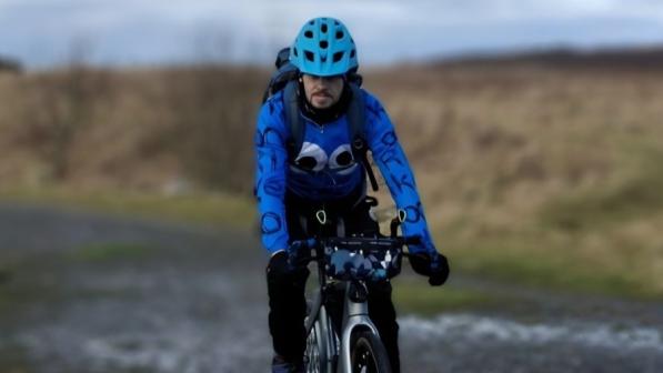 An adult cyclist wearing a bright blue helmet and a blue Cookie Monster-style jersey rides a grey bike along a muddy off-road path, with brown moorland and an overcast sky in the background.