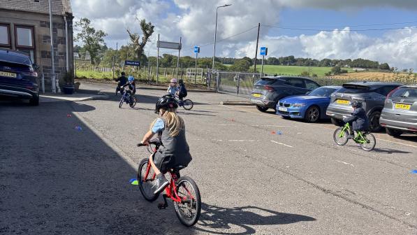 Children wearing helmets ride small bikes around coloured cones in a car park beside parked cars, with open countryside and a cloudy sky in the background.