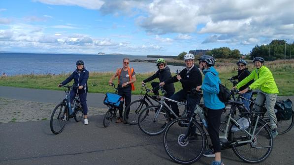 Eight helmeted cyclists pause with their bikes beside the sea during a group ride in Edinburgh.