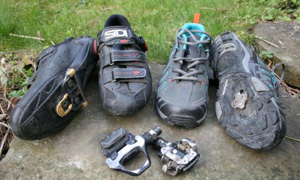 Four different types of clipless cycling shoes, with one lying on its side to show the cleat. In front are two types of clipless pedals