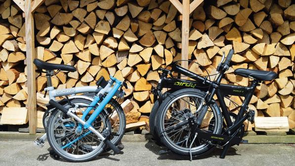 Two folding bikes are folded up in front of a pile of logs