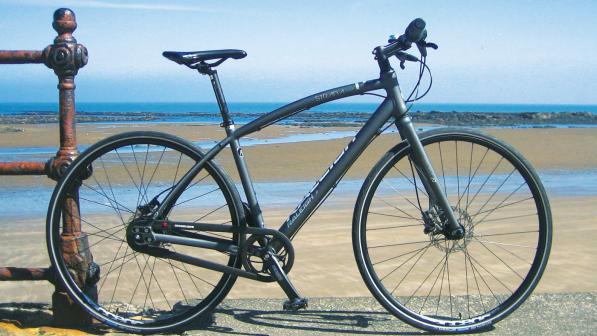A dark grey hybrid bike with flat handlebar, disc brakes and no mudguards. In the background is a beach with the tide far out