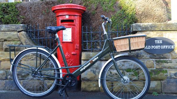 A dark green Elephant Bike. It's an upright, step-through bike with mudguards, long rear rack and a parcel rack with wicker basket at the front