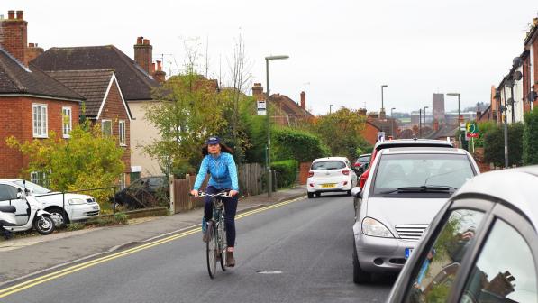 A woman is riding along a road in the centre of the lane so she can be seen. She is wearing a cap, blue jersey and blue leggings