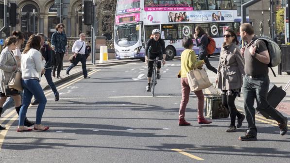 A woman is cycling on a busy city centre road. People are crossing in front of her and behind her is a bus