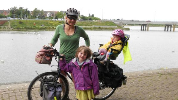 A woman in a green top is standing holding a bike. She has two little girls with her. One is standing in front of the bike and is wearing a purple coat and yellow dress, the other is in a child seat on the back of the bike