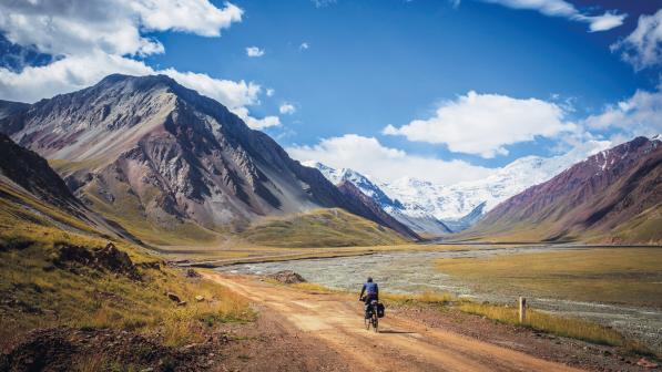 A person is riding a loaded touring bike in a valley with mountains in the background