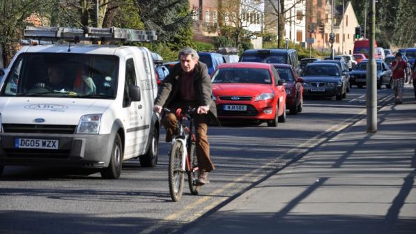 A man is cycling along a busy road with a line of traffic to his right