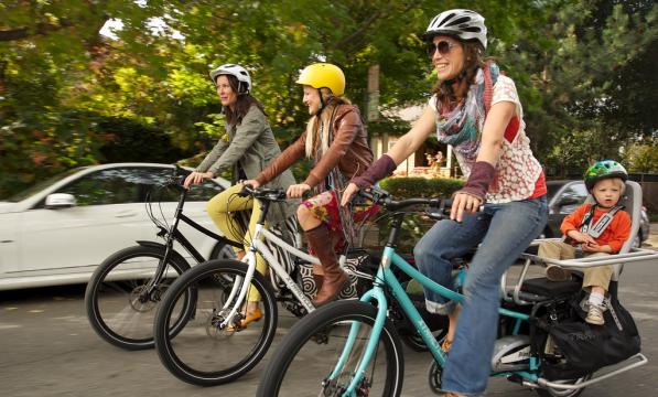 Three women are cycling along an urban road on e-cargo bikes. One has a child on the back of the bike. They are all wearing normal clothes and cycling helmets. There's a white car and trees in the background
